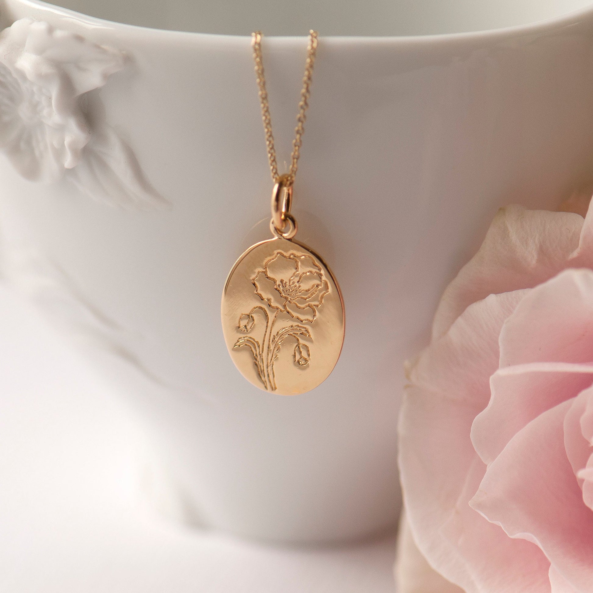 A gold-plated sterling silver necklace with an oval poppy pendant, displayed on a white surface with pink flowers in the background.