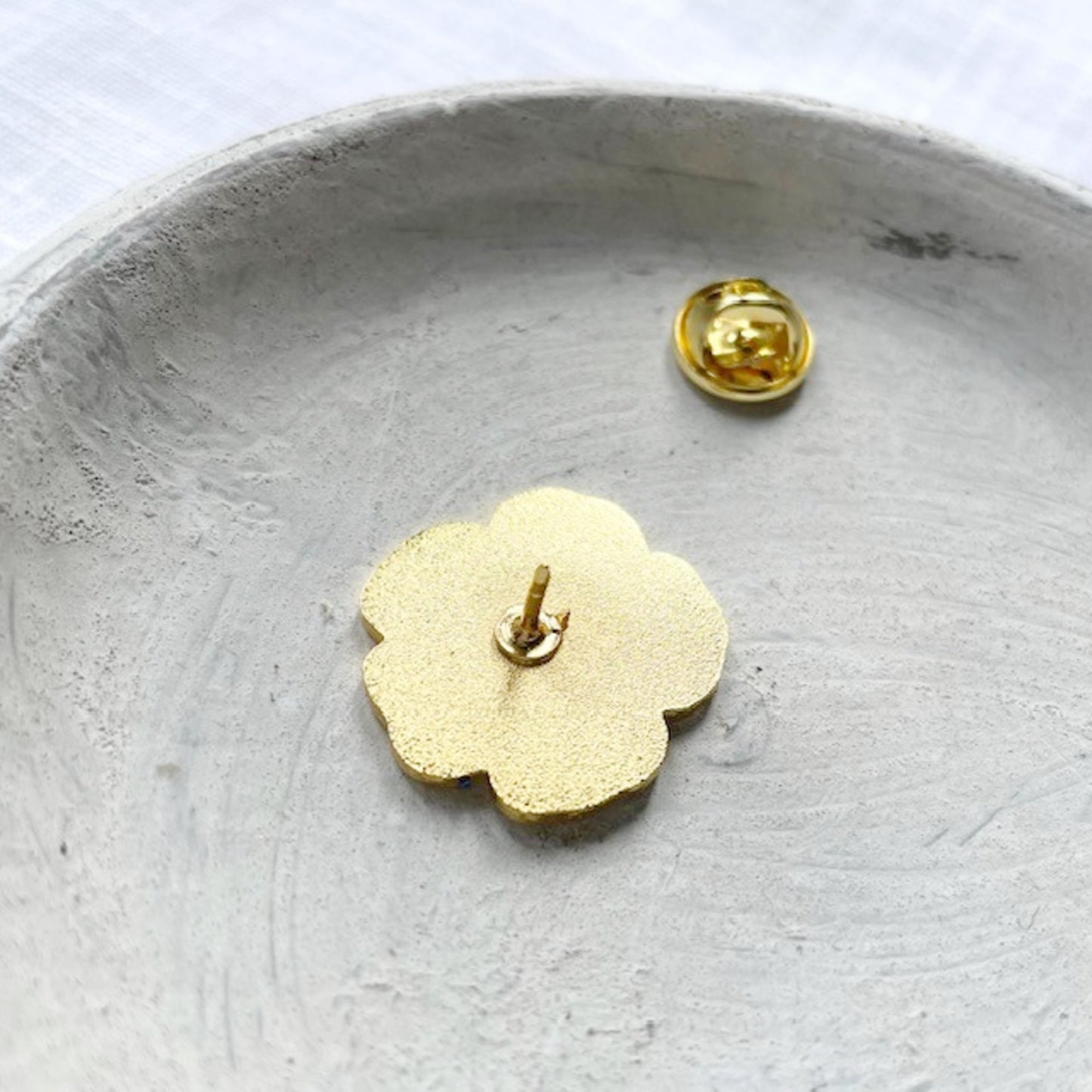 A small gold flower-shaped stud earring shown from the back, resting in a shallow gray dish with its matching gold ball back clip visible nearby.