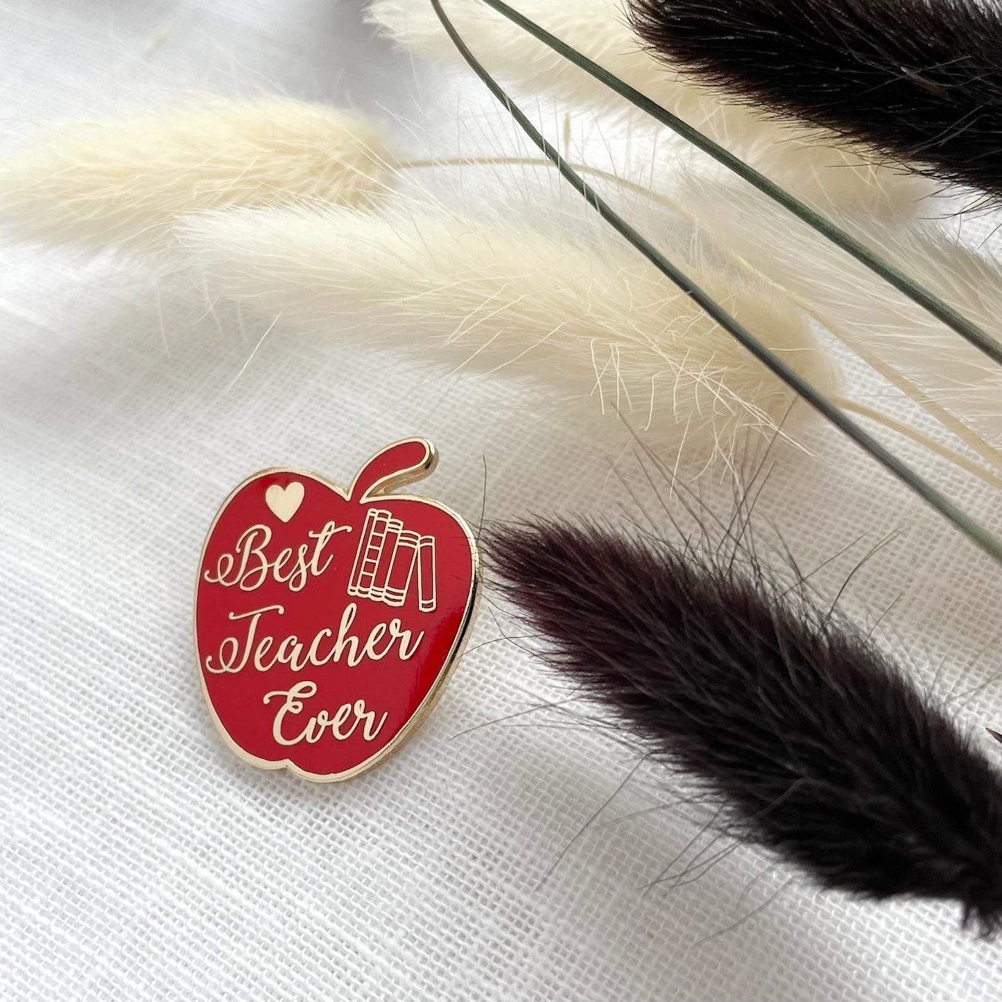 Red apple-shaped pin with 'Best Teacher Ever' text on a white surface close up shot showing the details of a pile of books and a love heart amongst the text