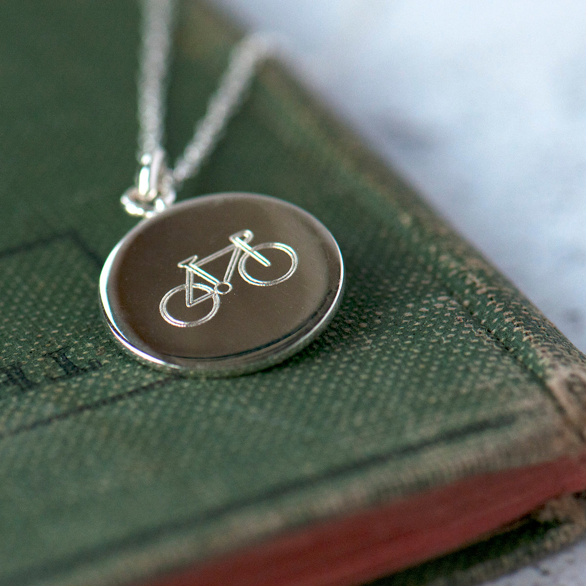 silver disc necklace wit the outline engraving of a bicycle resting on a book