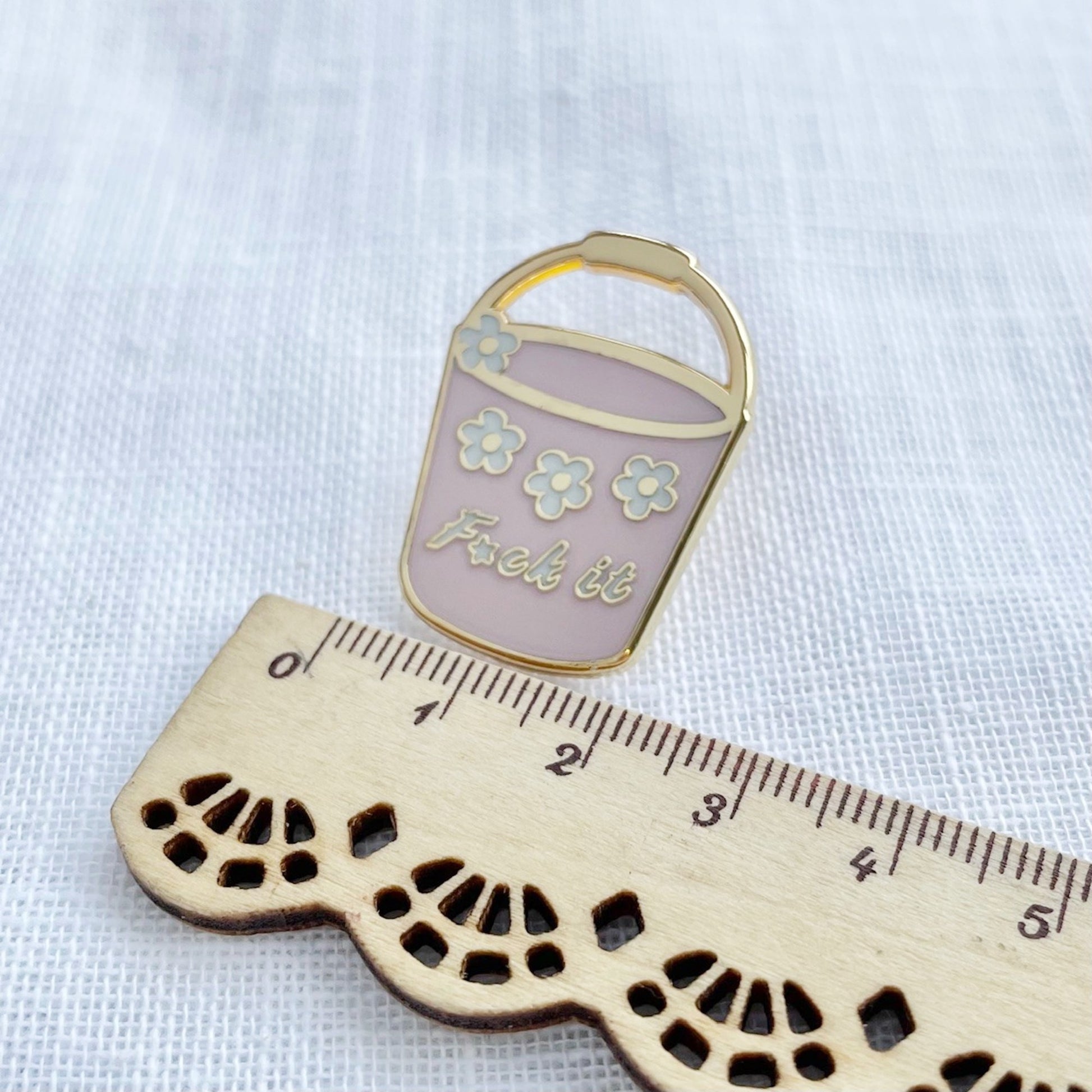 Enamel pin of a pink bucket with flowers and 'F*ck it' text, next to a wooden ruler on a white background