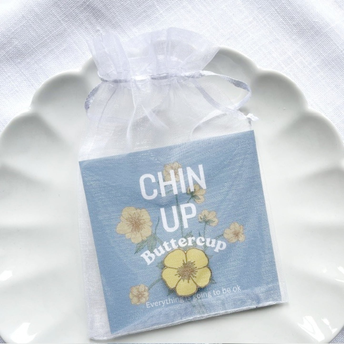 A small square card or sticker package tied in a sheer white organza drawstring bag, placed on a white scalloped plate. The package has a light blue background with yellow flower illustrations and the words “CHIN UP Buttercup” printed on it.