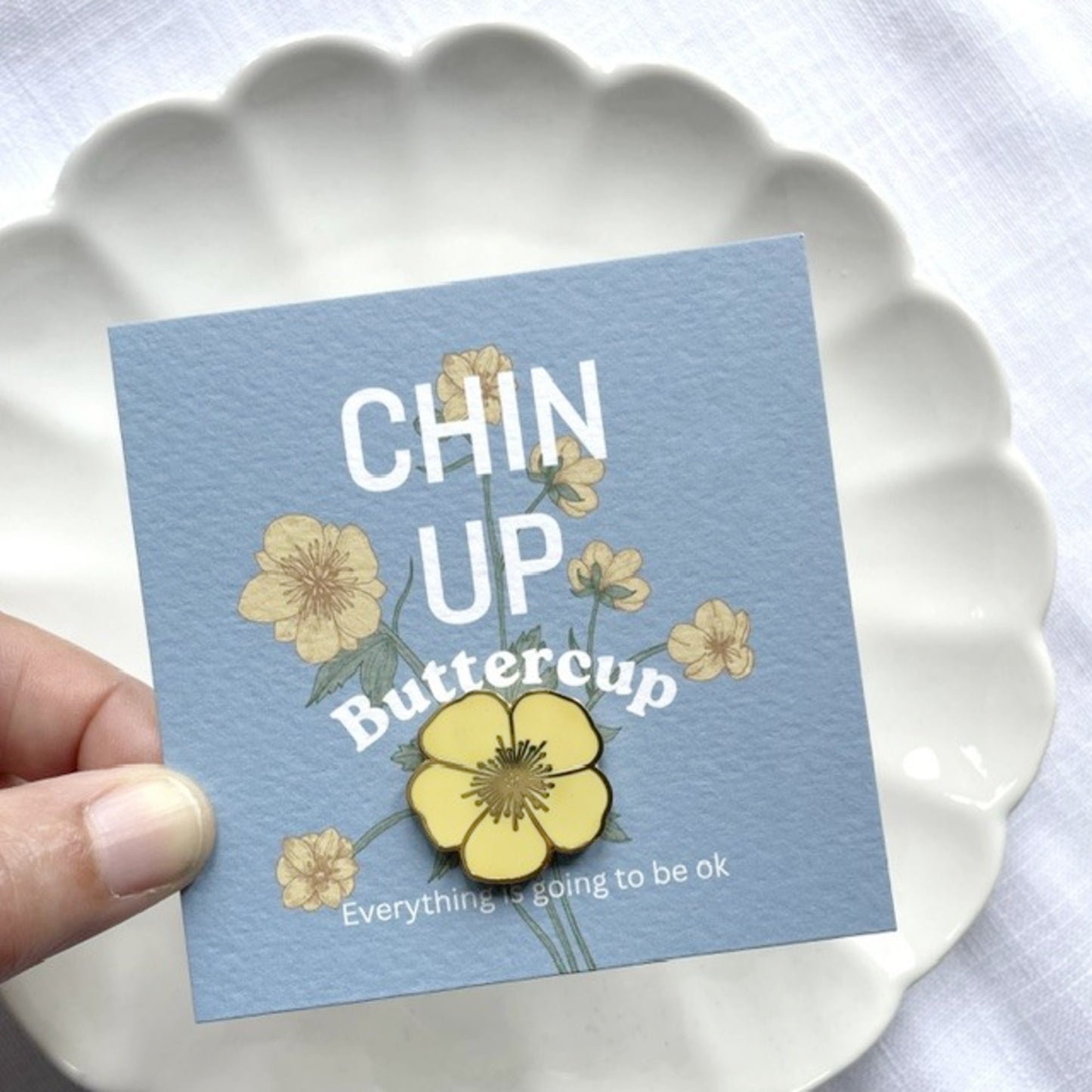 A hand holds a square light blue card with the phrase “CHIN UP Buttercup” and small yellow flower illustrations, positioned over a white scalloped plate.