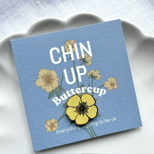A square blue buttercup gift card with the text 'CHIN UP Buttercup' and smaller text 'Everything is going to be ok,' decorated with yellow buttercup flowers, shown on a white scalloped dish against a light textured background.