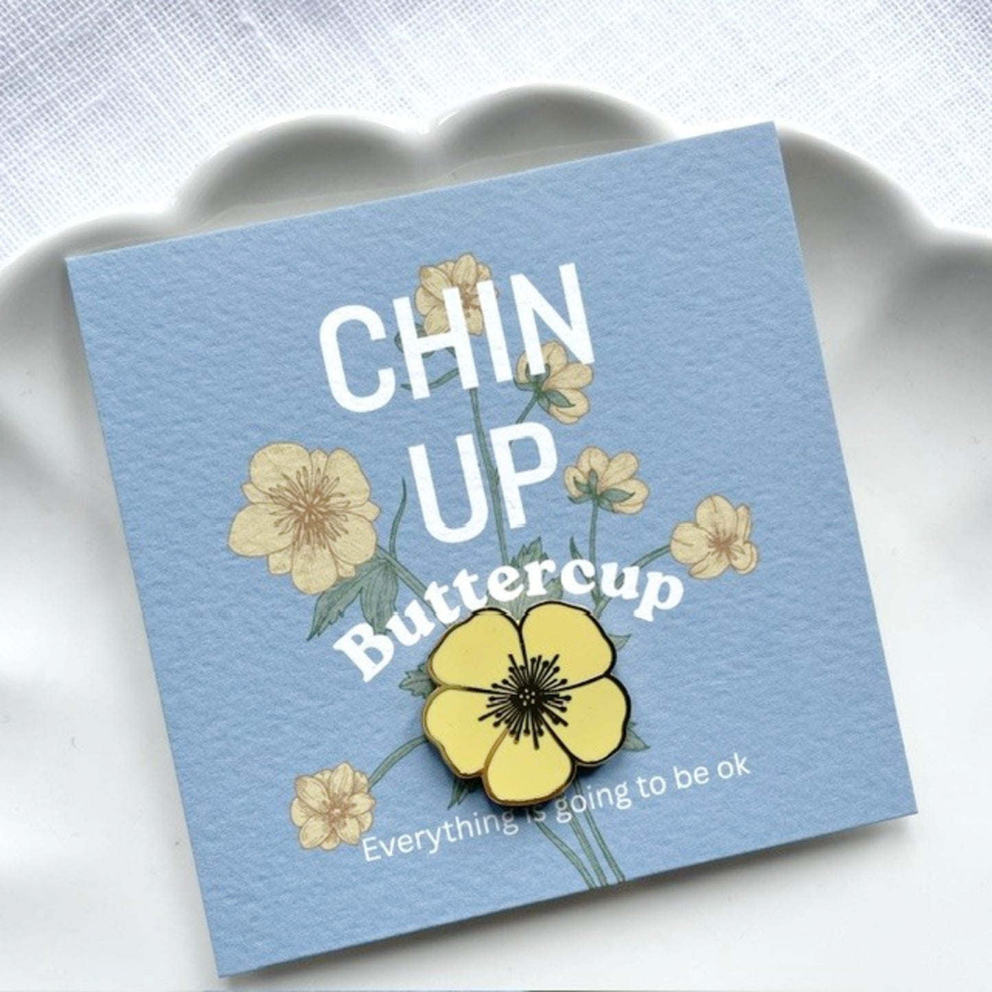 A square blue gift card featuring the text 'CHIN UP Buttercup' and 'Everything is going to be ok' with yellow flower illustrations, photographed and a sunshine yellow buttercup pin.