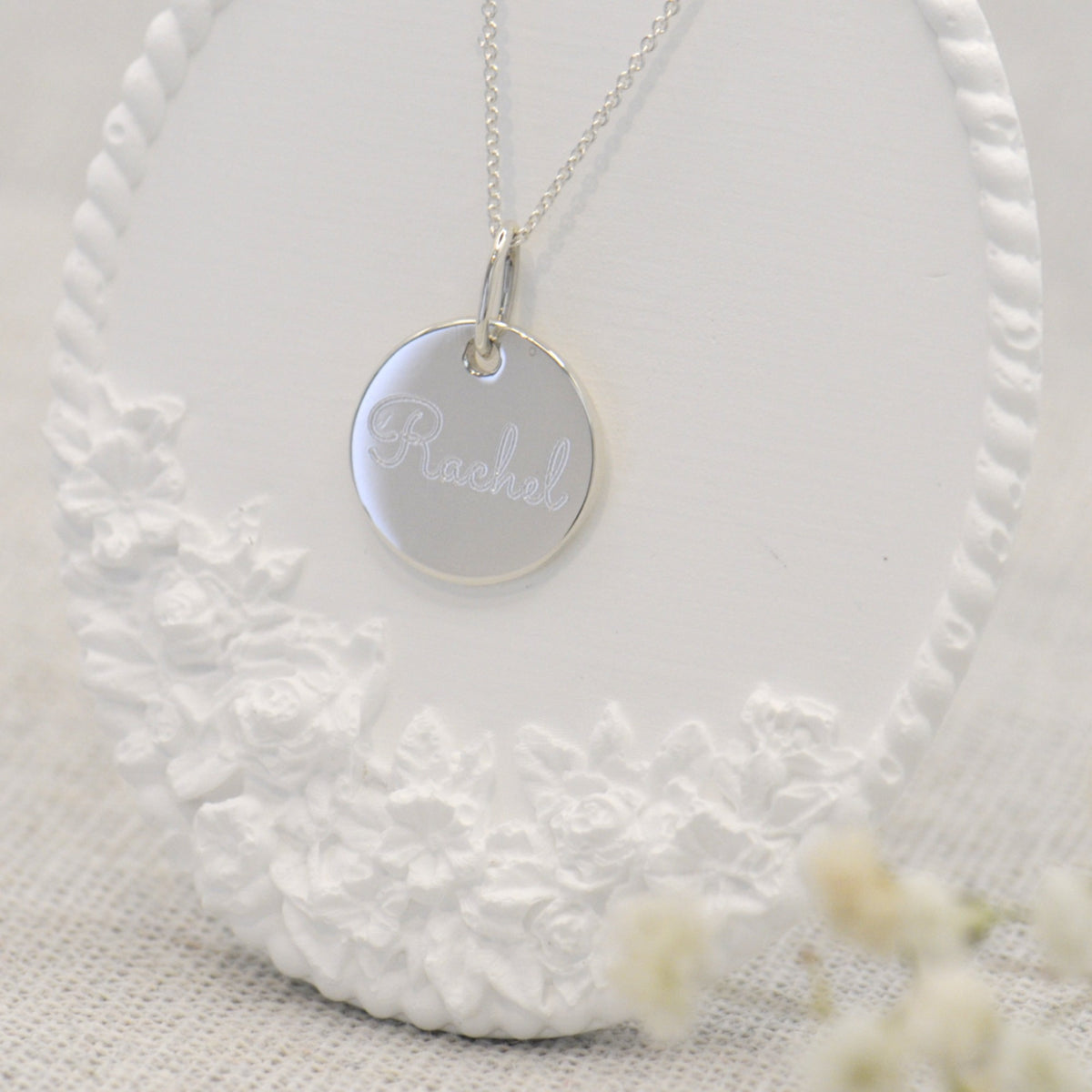 A silver round pendant necklace engraved with the name 'Rachel', displayed on a white floral stand.
