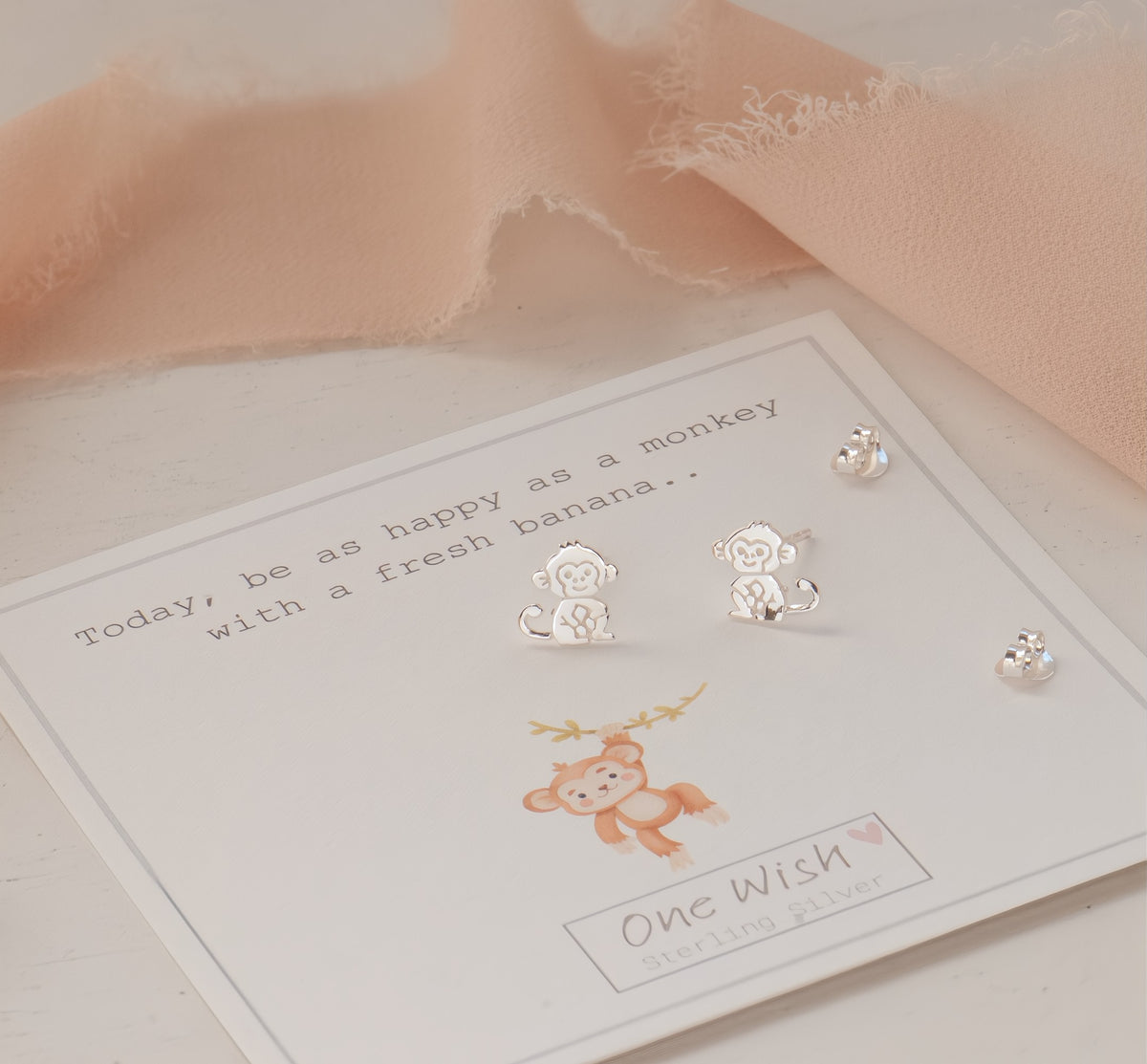 Sterling silver monkey stud earrings displayed on a white card labeled 'One Wish Sterling Silver', with a peach fabric accent in the background.