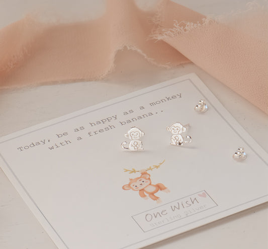 Sterling silver monkey stud earrings displayed on a white card labeled 'One Wish Sterling Silver', with a peach fabric accent in the background.
