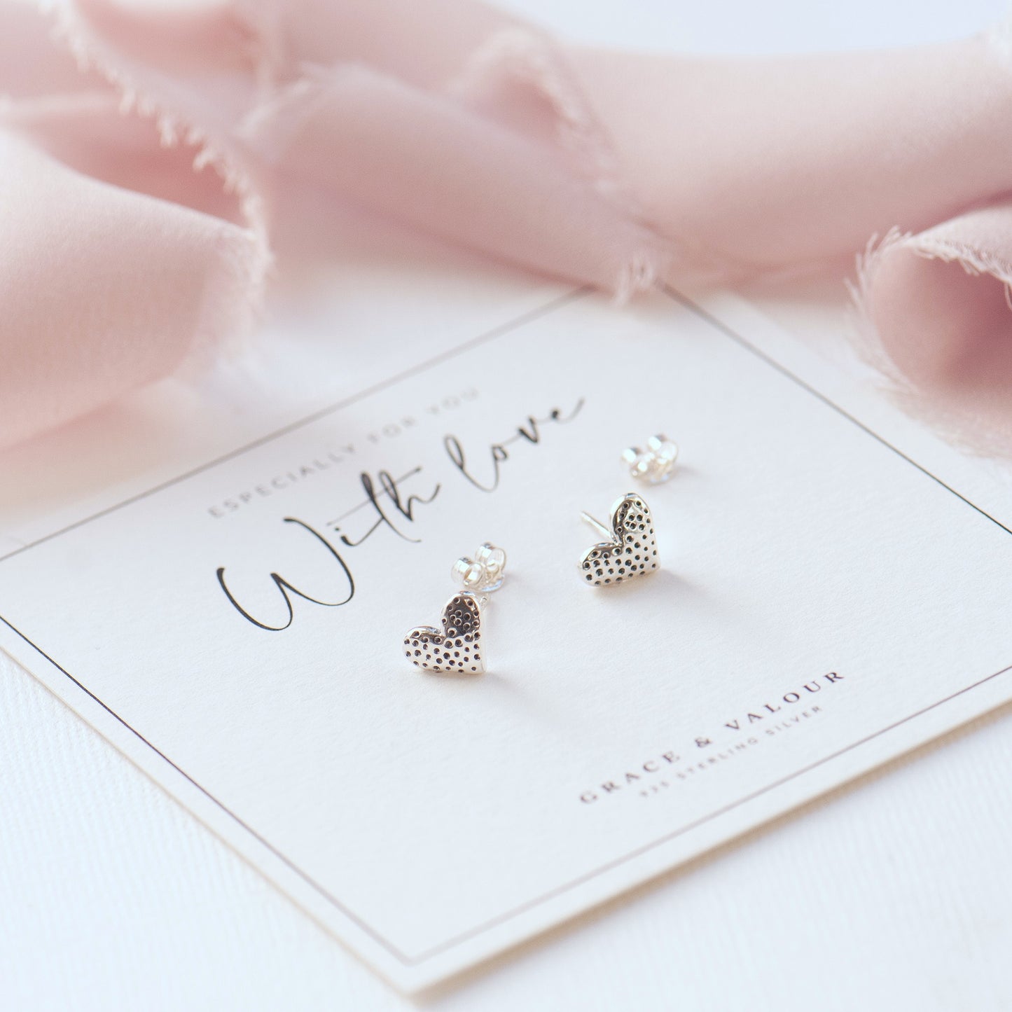 A pair of silver heart stud earrings with a textured, dotted pattern, displayed on a white card that reads 'With love' and 'Grace & Valour'. The card is styled with soft pink fabric in the background.