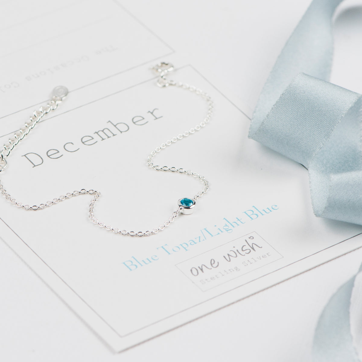 A delicate silver chain bracelet with a small round light blue topaz gemstone, displayed on its white presentation card next to a light blue satin ribbon.