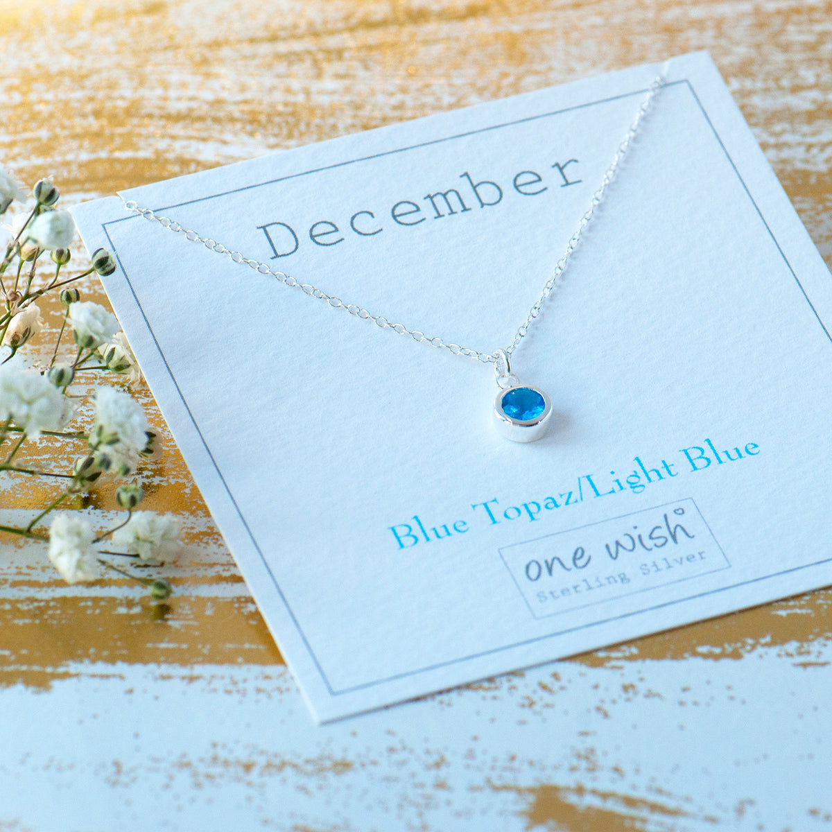 A sterling silver necklace with a round light blue topaz pendant, displayed on a white card labeled 'December' and 'Blue Topaz/Light Blue' by the brand 'one wish', set on a rustic wooden surface with baby's breath flowers.