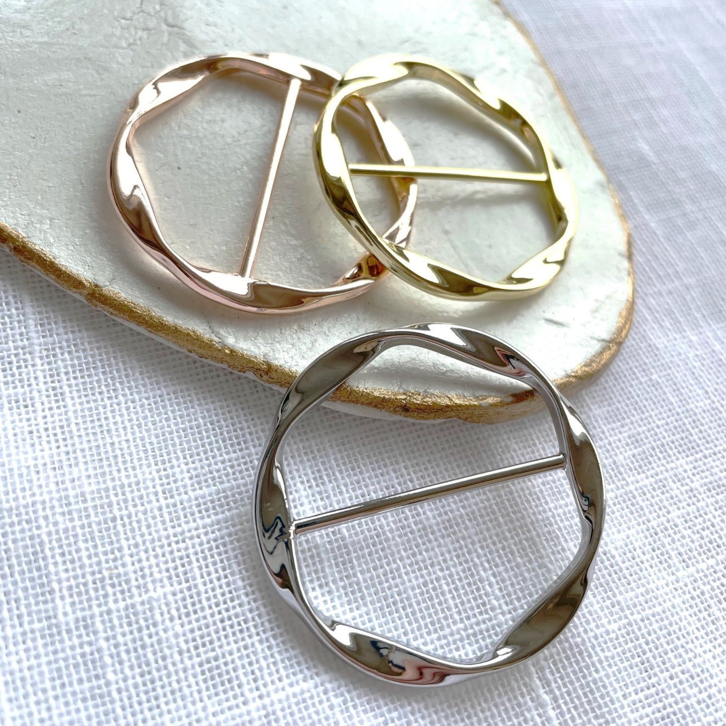 Three metal round scarf holders in gold, rose gold, and silver on a textured white surface.