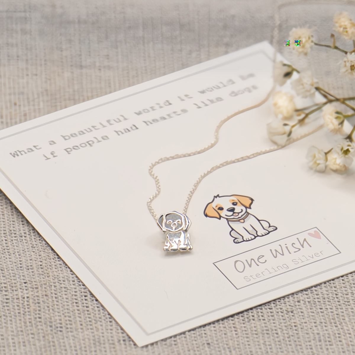 A sterling silver dog pendant necklace displayed on a white gift card with a printed dog illustration and the text 'One Wish Sterling Silver,' styled on a textured beige fabric with small white flowers.
