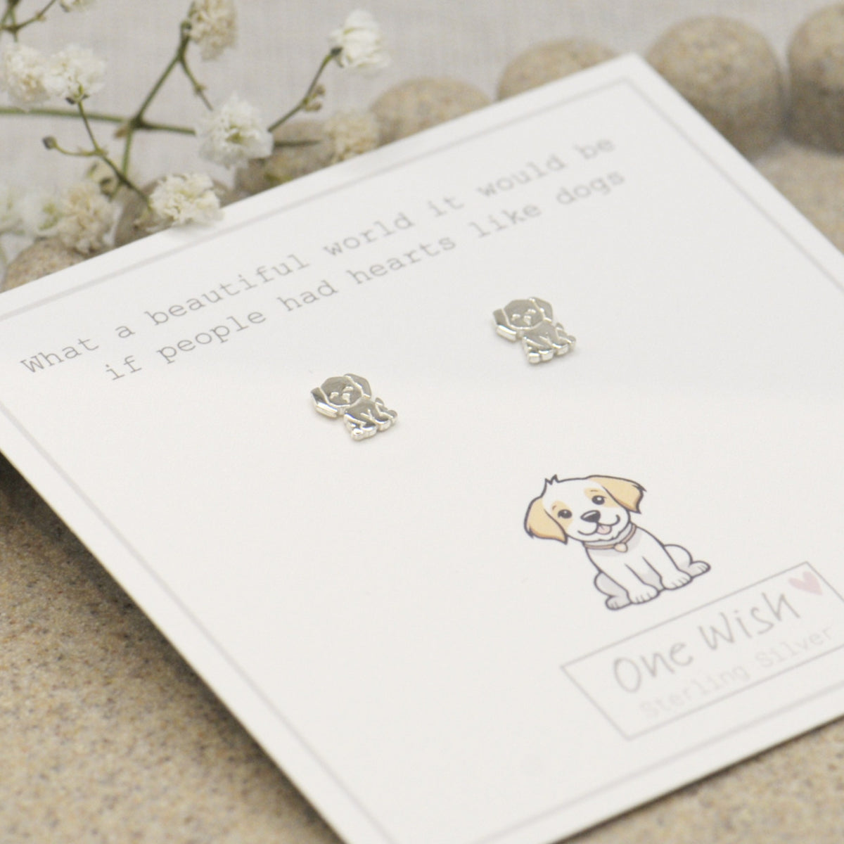 A pair of small silver stud earrings shaped like dogs, displayed on a white gift card with printed text and a puppy illustration. The card is photographed on a soft neutral surface with small white flowers in the background.