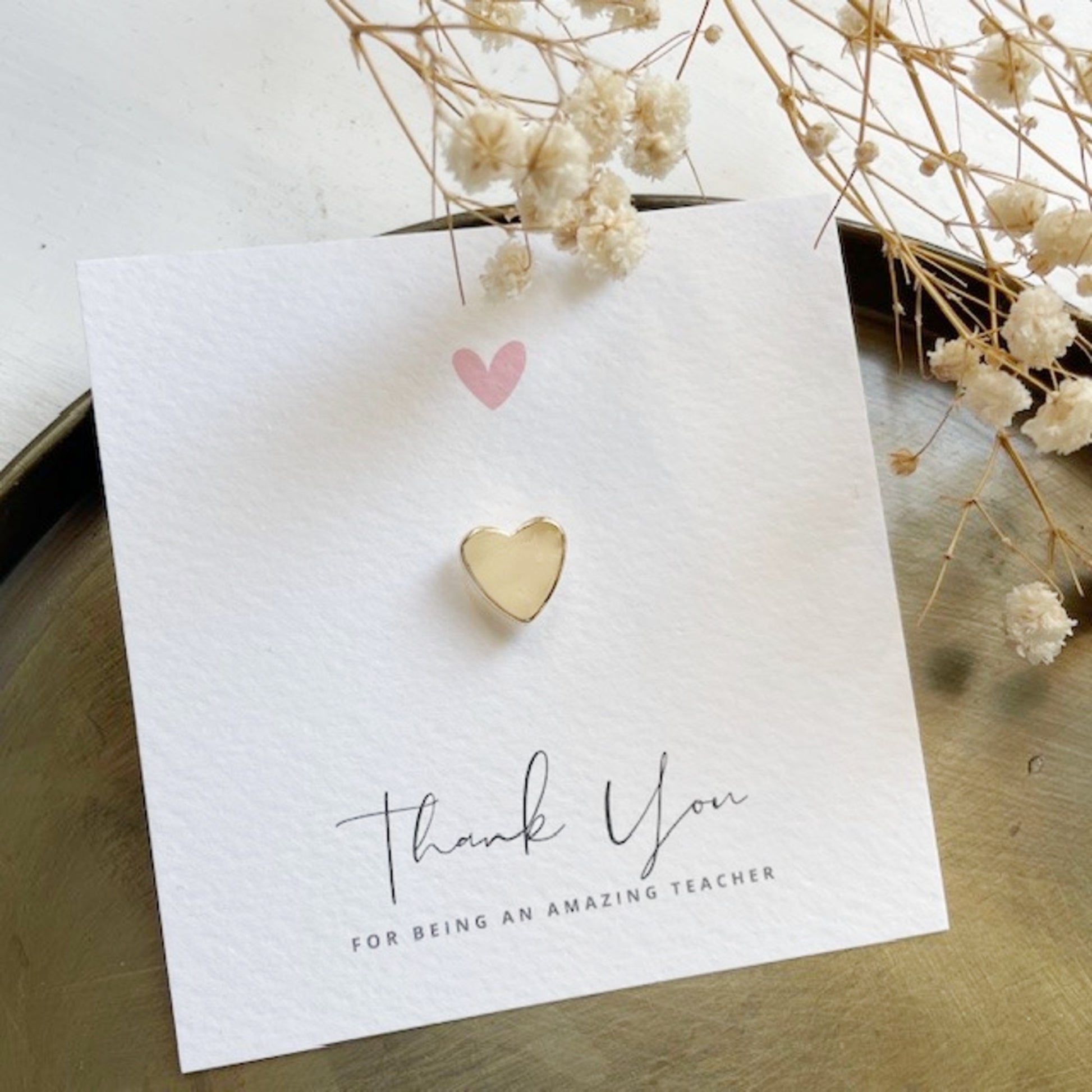 Heart-shaped gold metal pin on a 'Thank You for Being an Amazing Teacher' card with dried flowers.