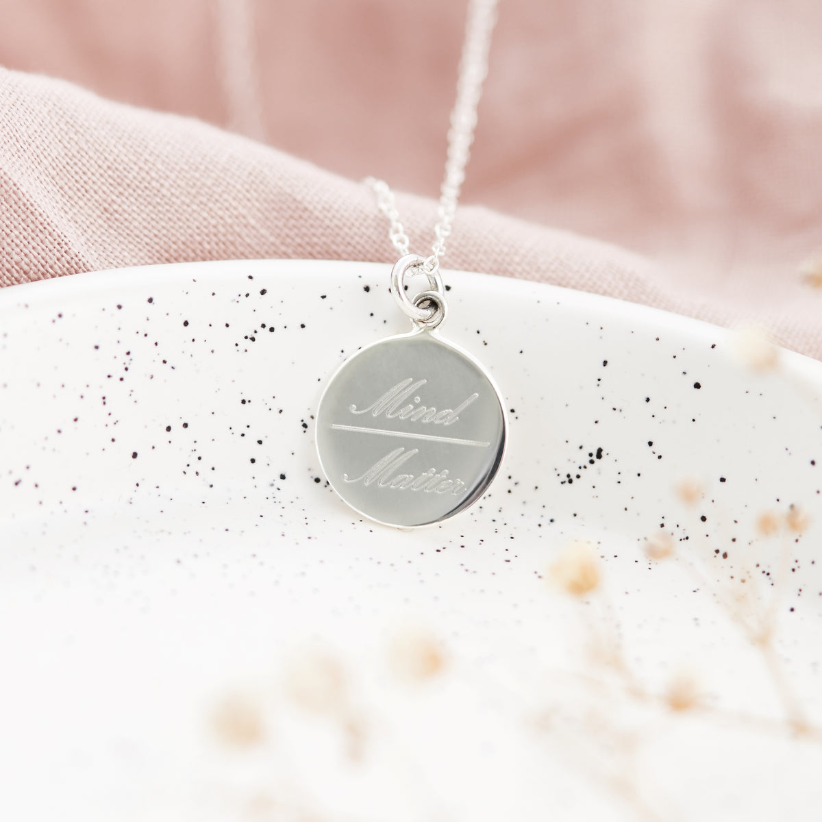 A silver round pendant necklace engraved with the words 'Mind Matters' in script and serif fonts, resting on a white speckled ceramic plate with a soft pink fabric background.