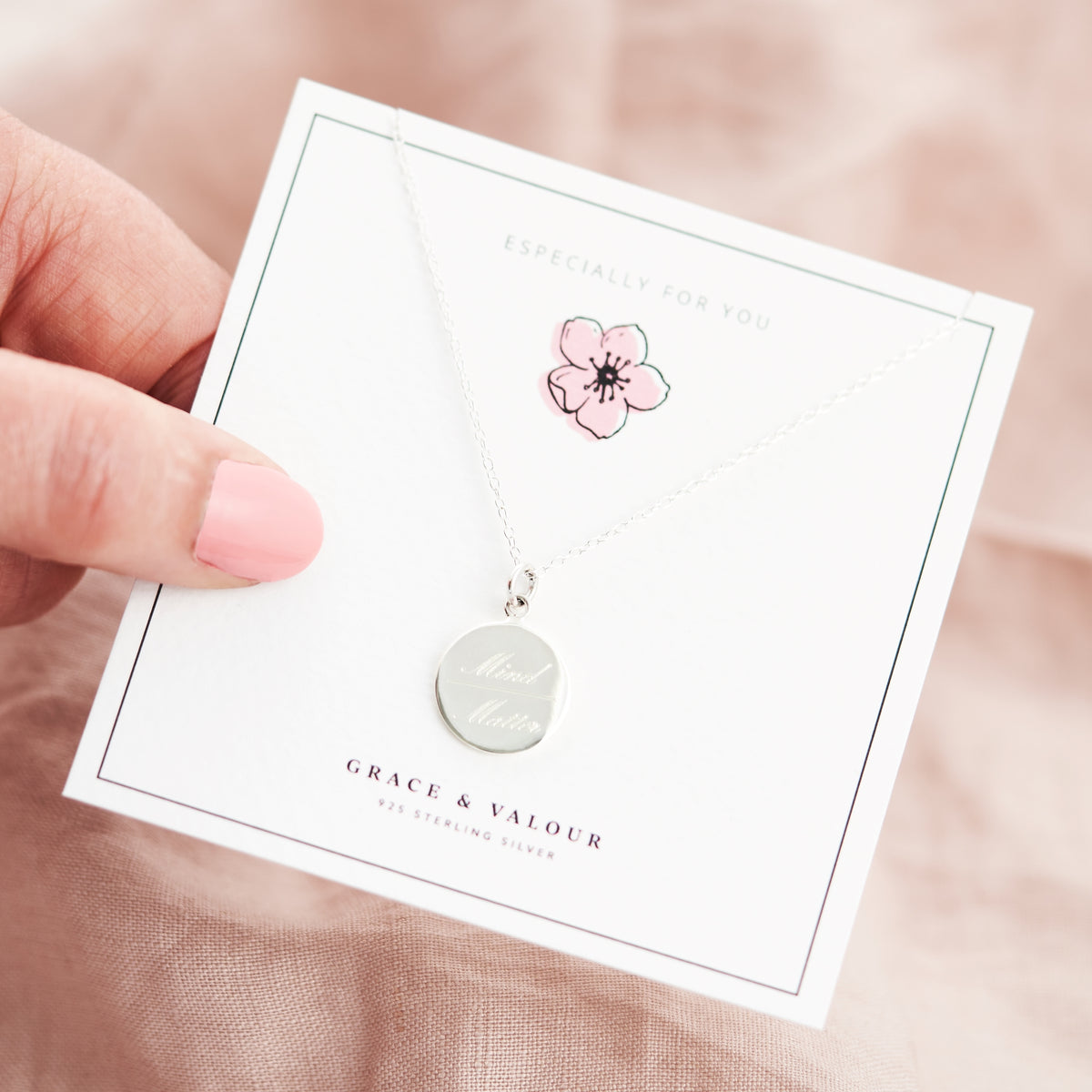 A silver necklace with a round engraved disc pendant is displayed on a white jewelry card held by a hand. The card features a pink cherry blossom illustration and the brand name Grace & Valour, set against a soft pink fabric background.