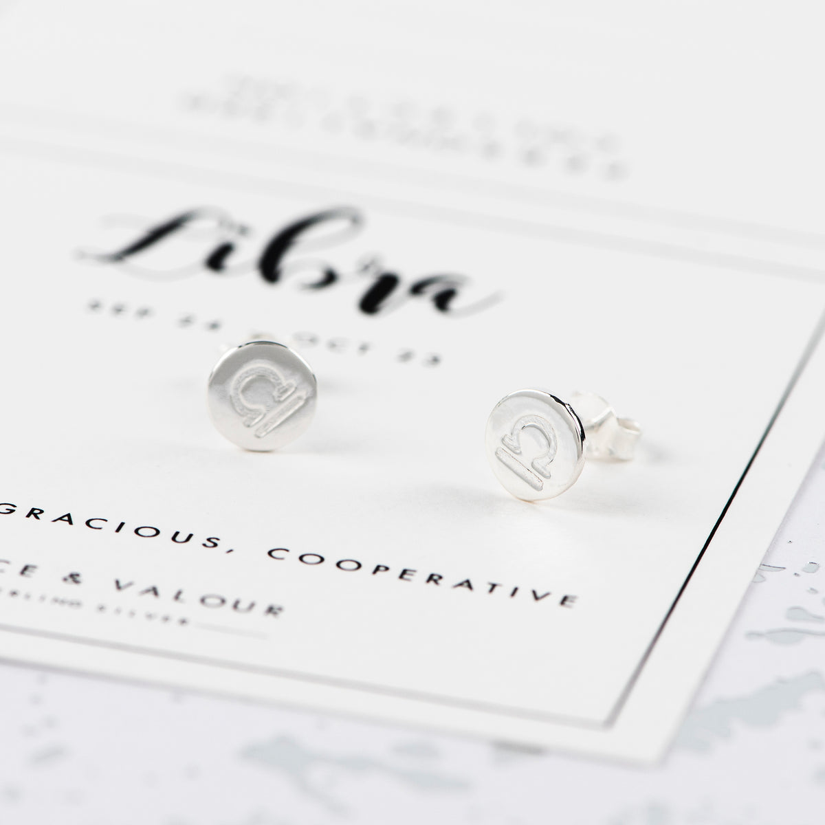 A pair of round silver stud earrings with an engraved symbol for the star sign libra on a white card printed with the word 'Libra'.