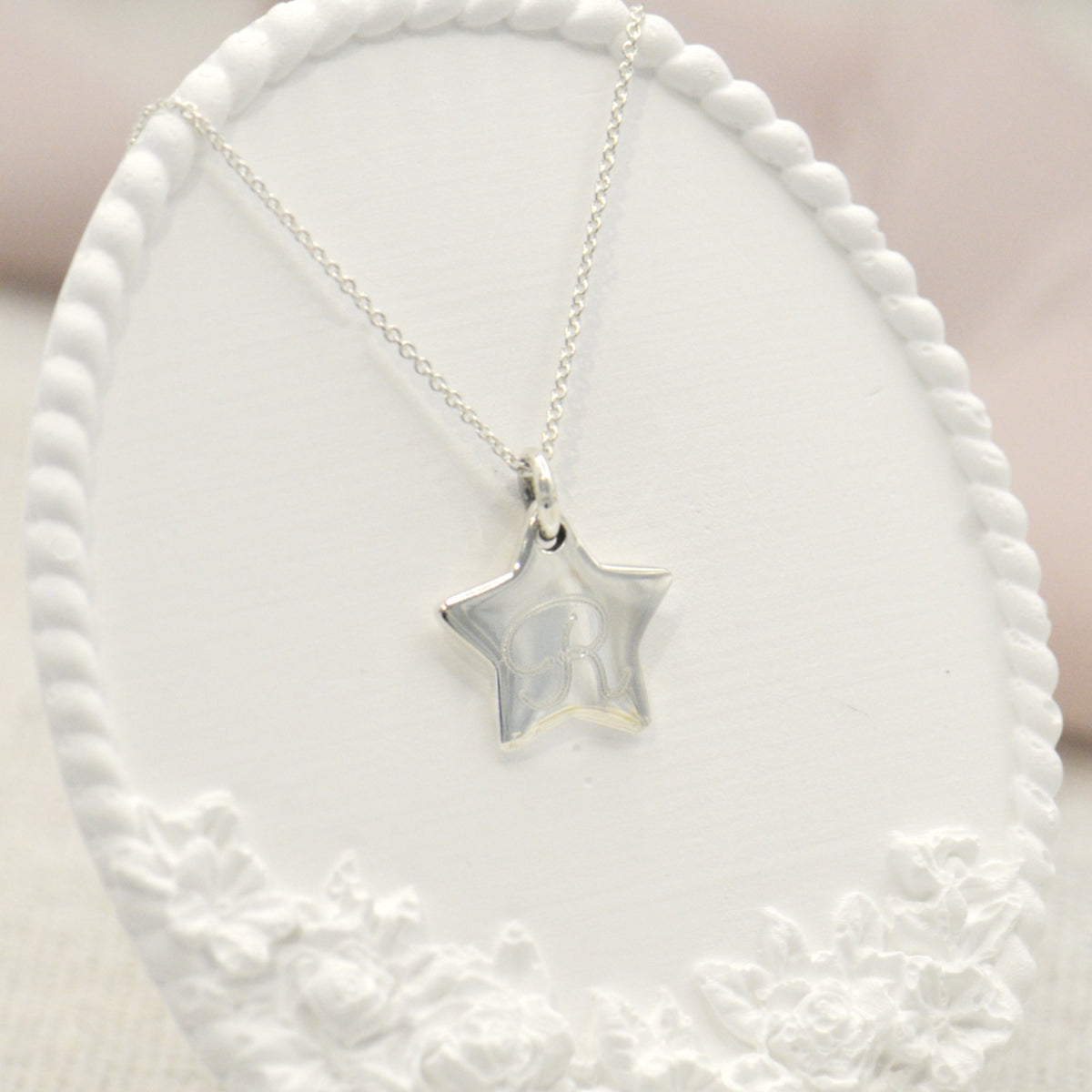 A silver star pendant necklace with a delicate chain, displayed on a white oval jewelry bust with a floral lace border.
