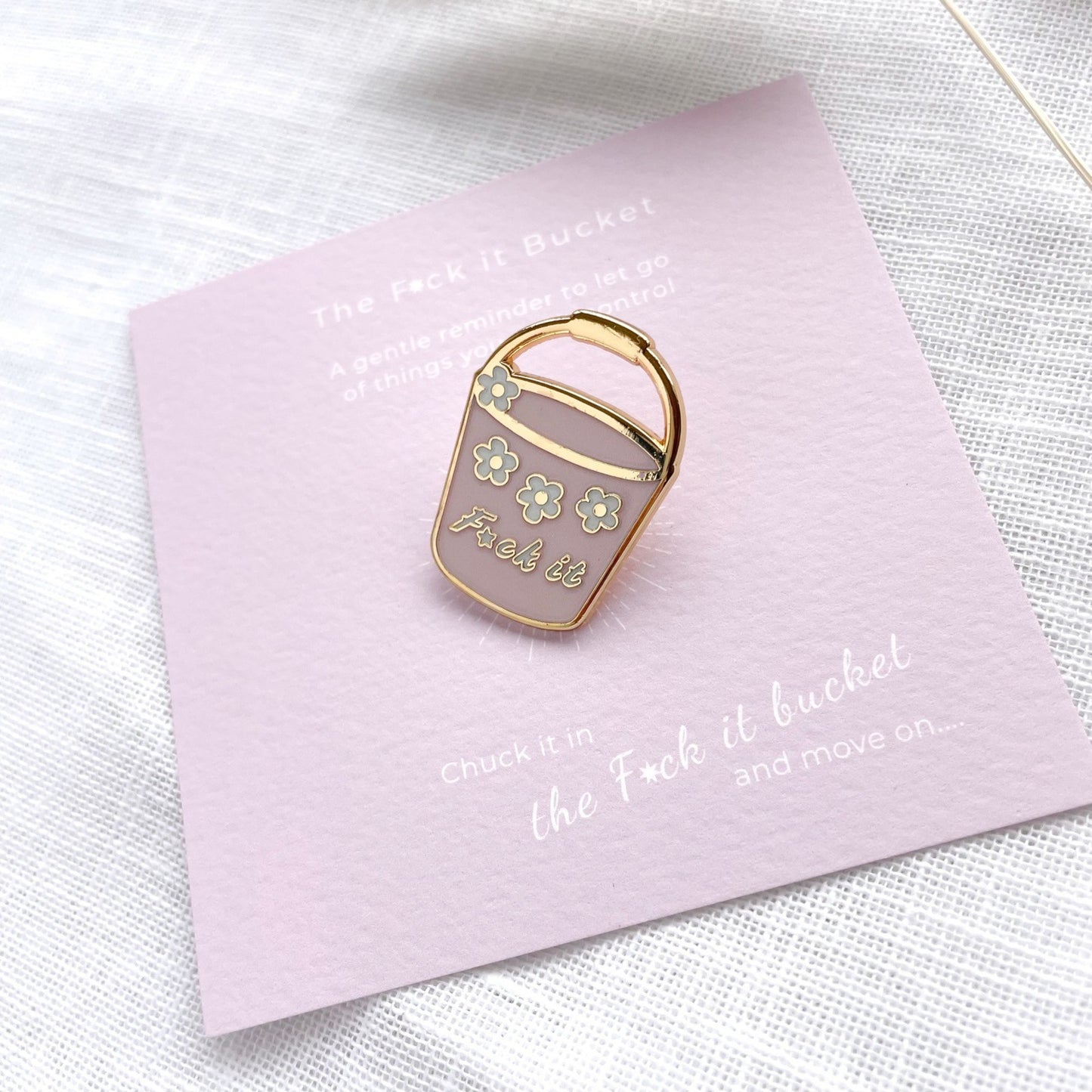 Enamel pin shaped like a bucket with floral design on a pink card with text.