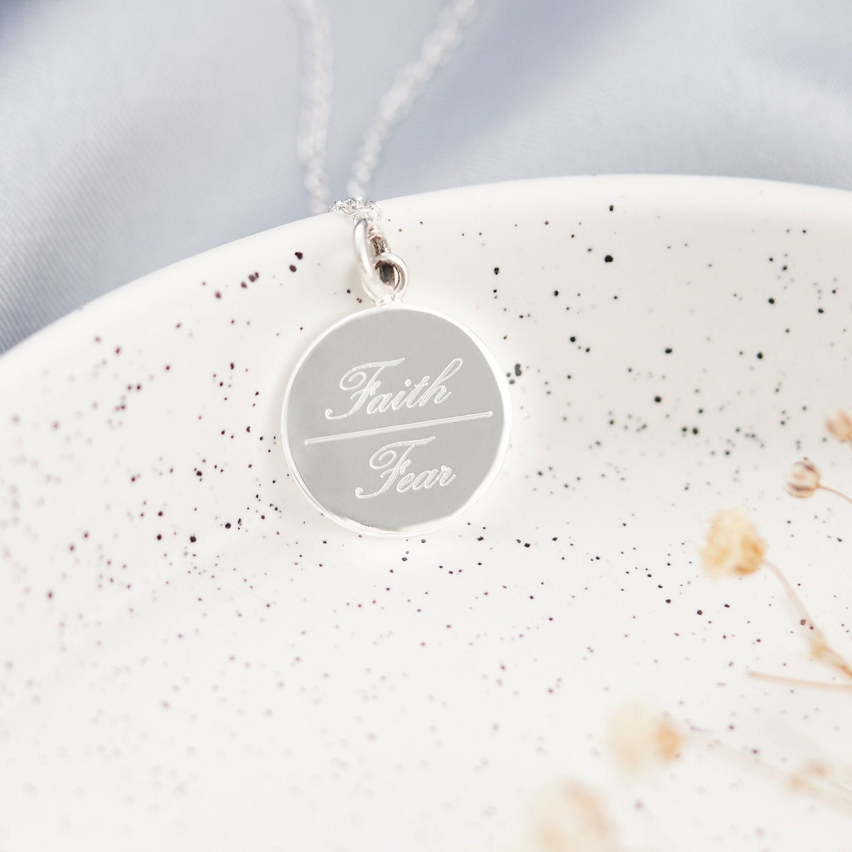silver disc necklace on a speckled background engraved with the word faith over the word fear