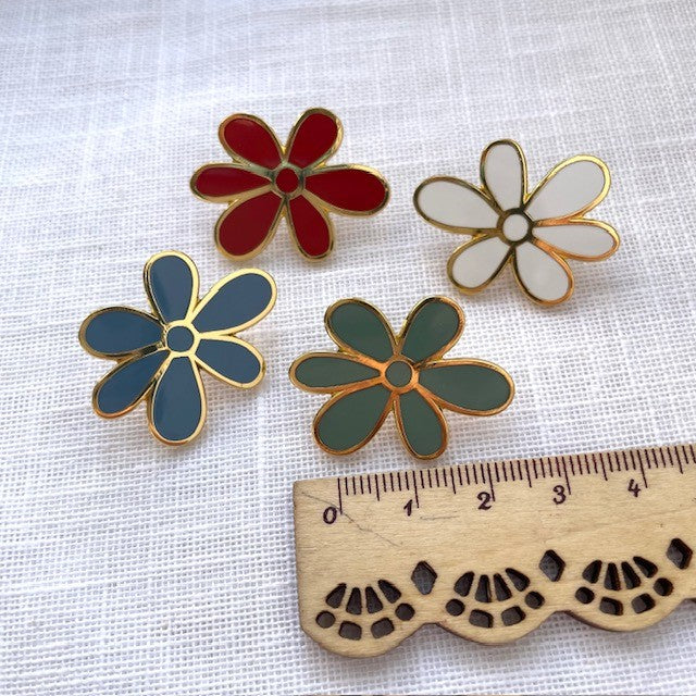 Four colorful flower-shaped pins on a white fabric background with a wooden ruler for scale.