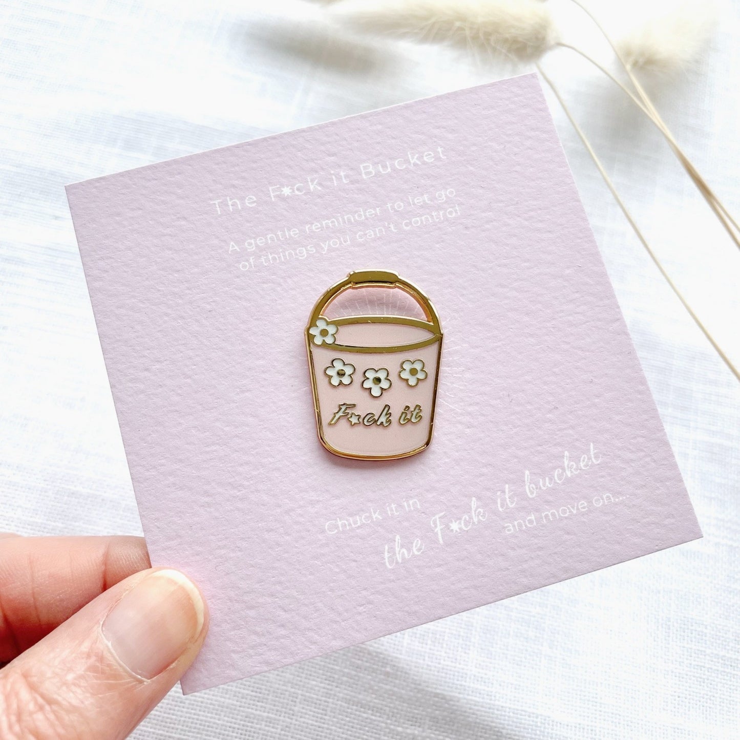 Enamel pin of a f*ck it bucket with flowers on a card held by a hand.