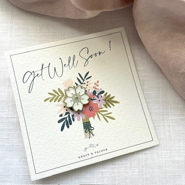 Get Well Soon card with bouquet of flowers gift card and a white enamel flower pin among the flowers