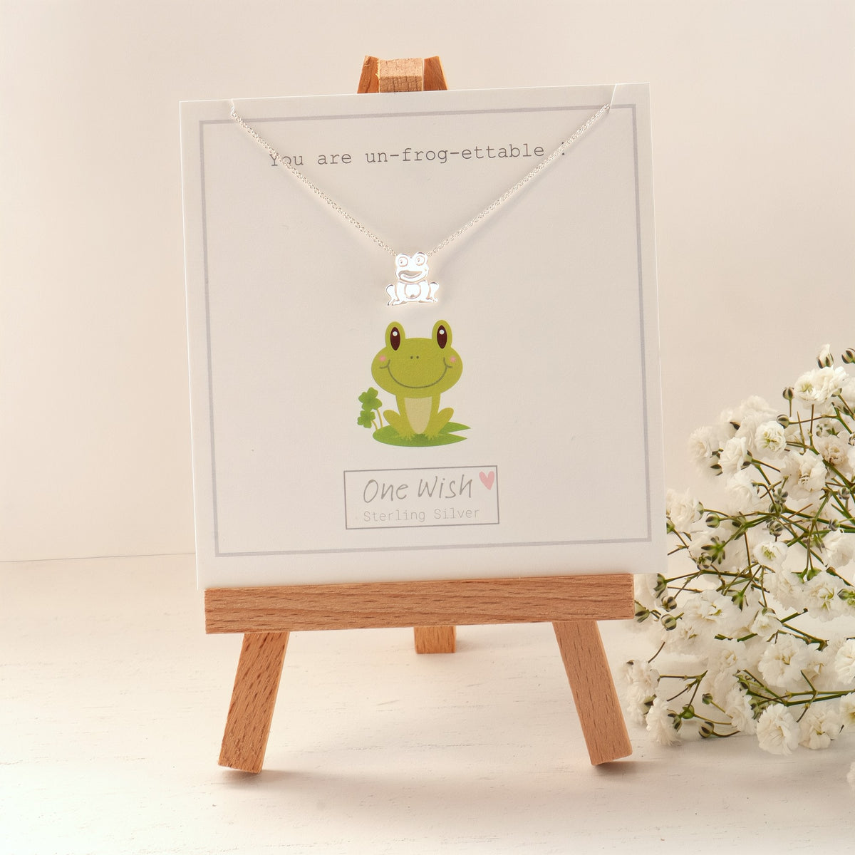 A pair of silver stud earrings in the shape of a frog displayed on a white card with a quote that reads 'You are Un-Frog-ettable', presented on a wooden easel with a white flower in the background.