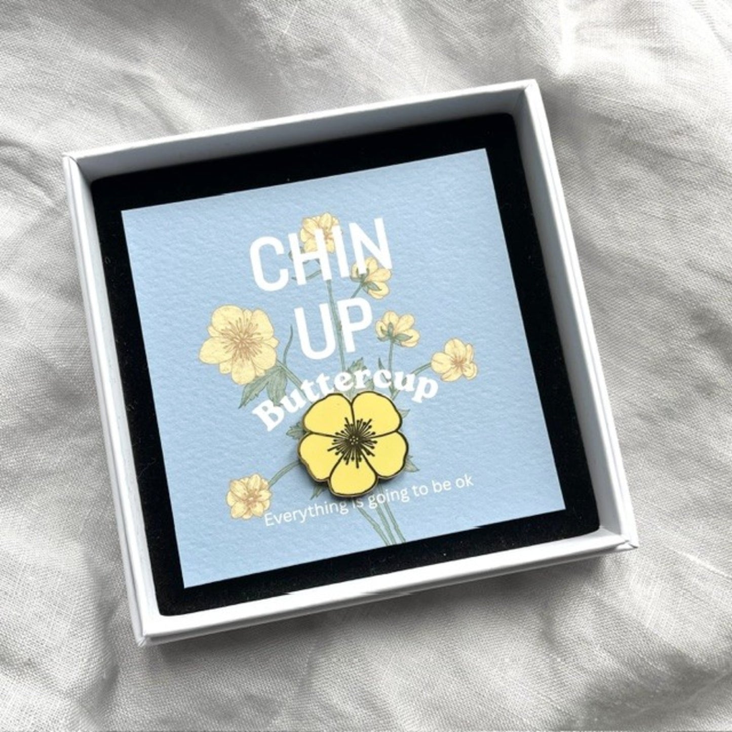 A square white gift box with a black inner border holds a light blue card printed with the words “CHIN UP Buttercup” and yellow flower illustrations. The box is photographed on a soft white fabric background.