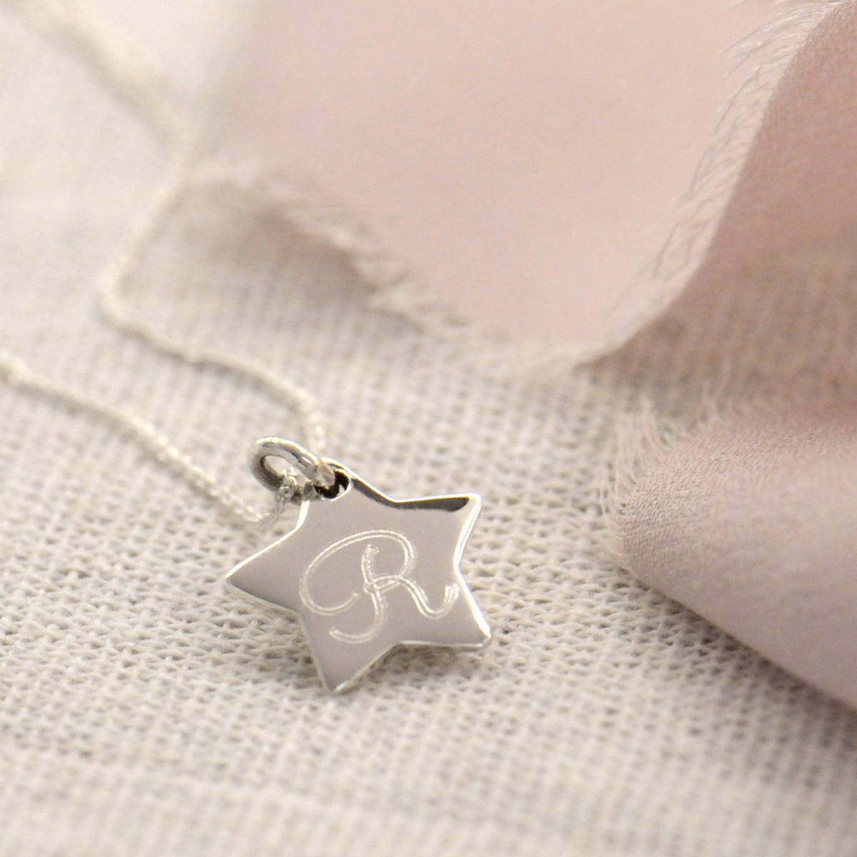 A silver star-shaped pendant necklace with an engraved letter 'R', resting on a textured beige fabric next to soft pink ribbon.