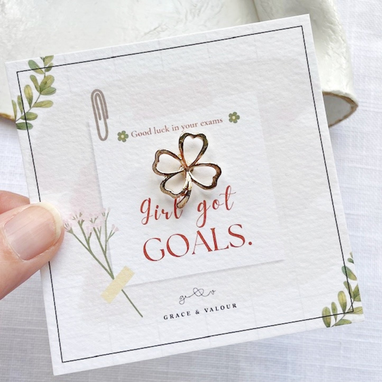 Gold four-leaf clover charm on a card with 'Girl got goals' text, held by a hand.