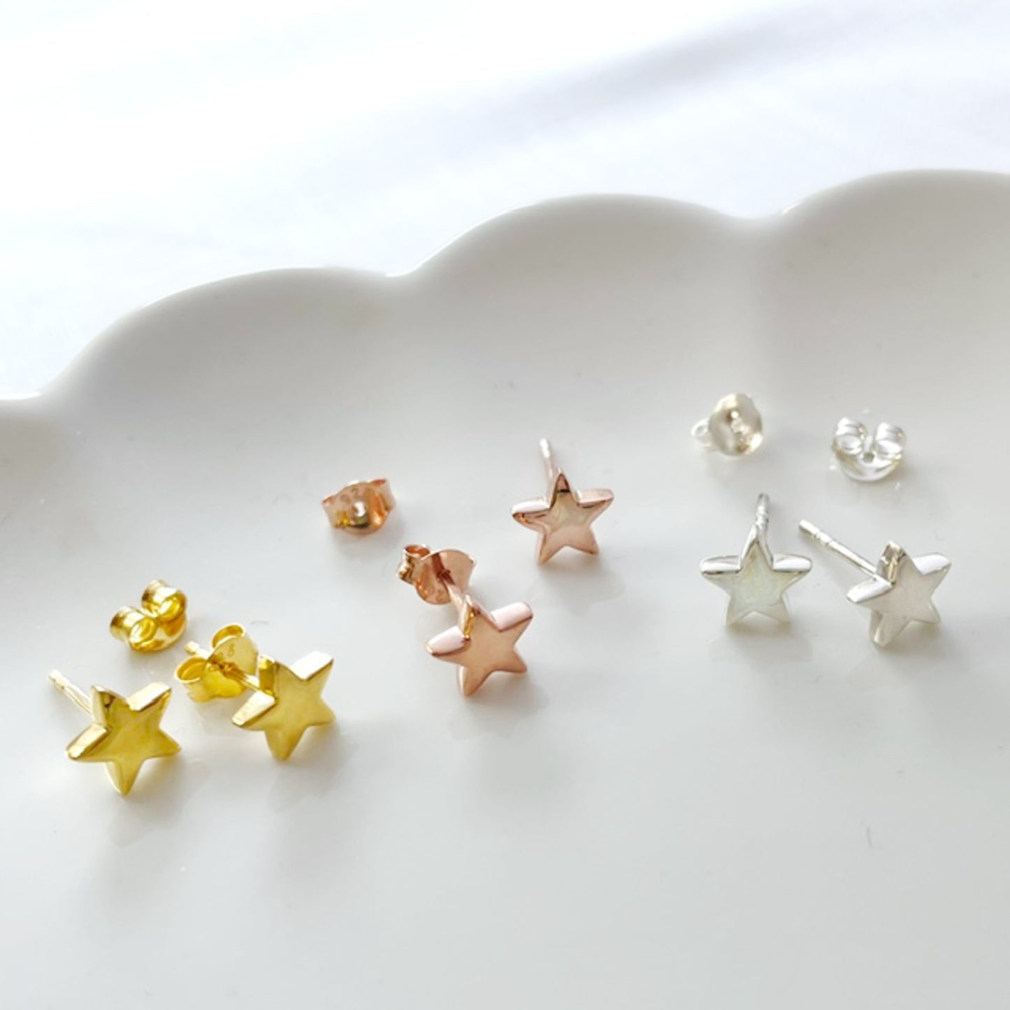 A set of small star-shaped stud earrings in yellow, rose gold, and silver, arranged on a white scalloped dish.