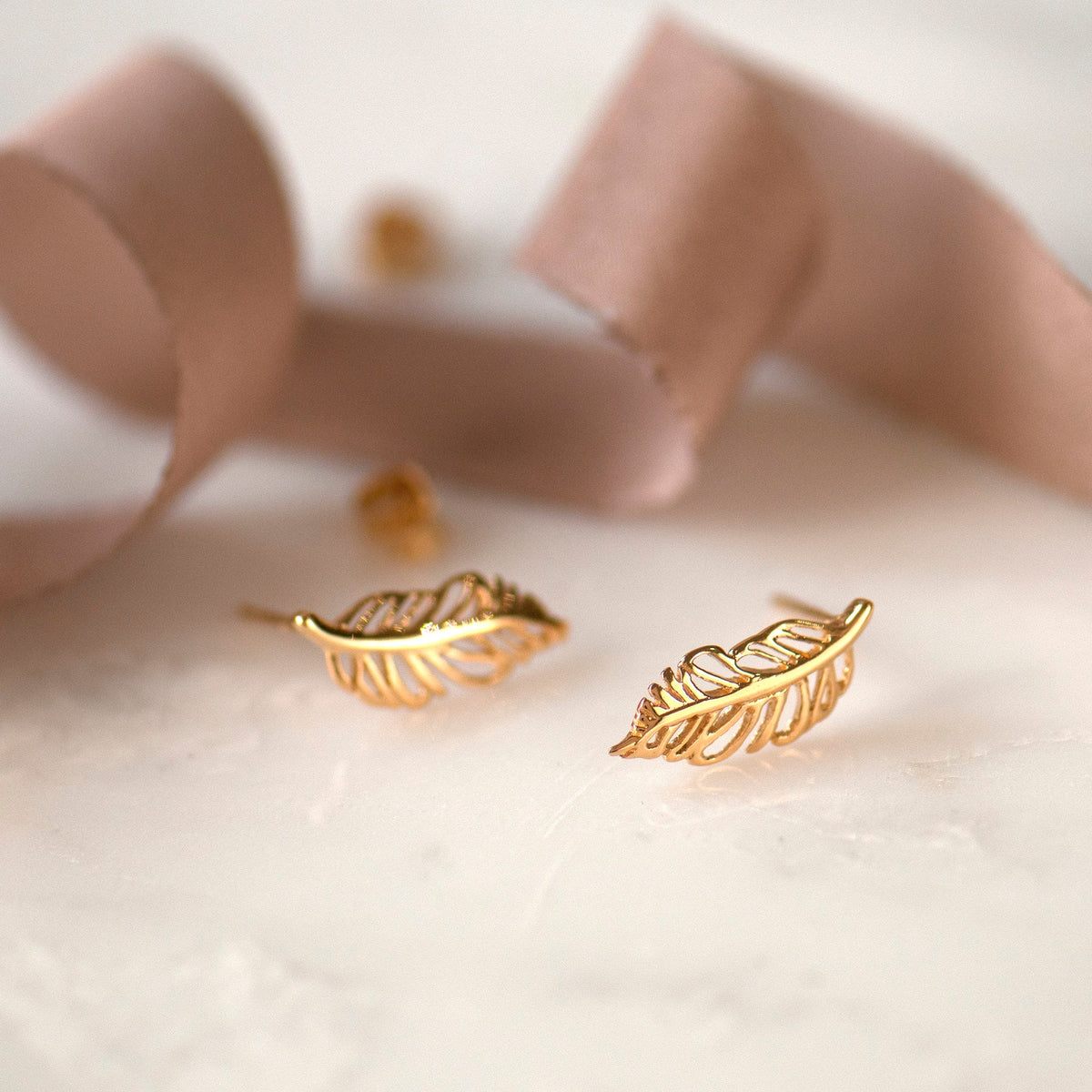 Gold-colored feather-shaped stud earrings on a white marble surface, with a pink ribbon and a silver butterfly back.