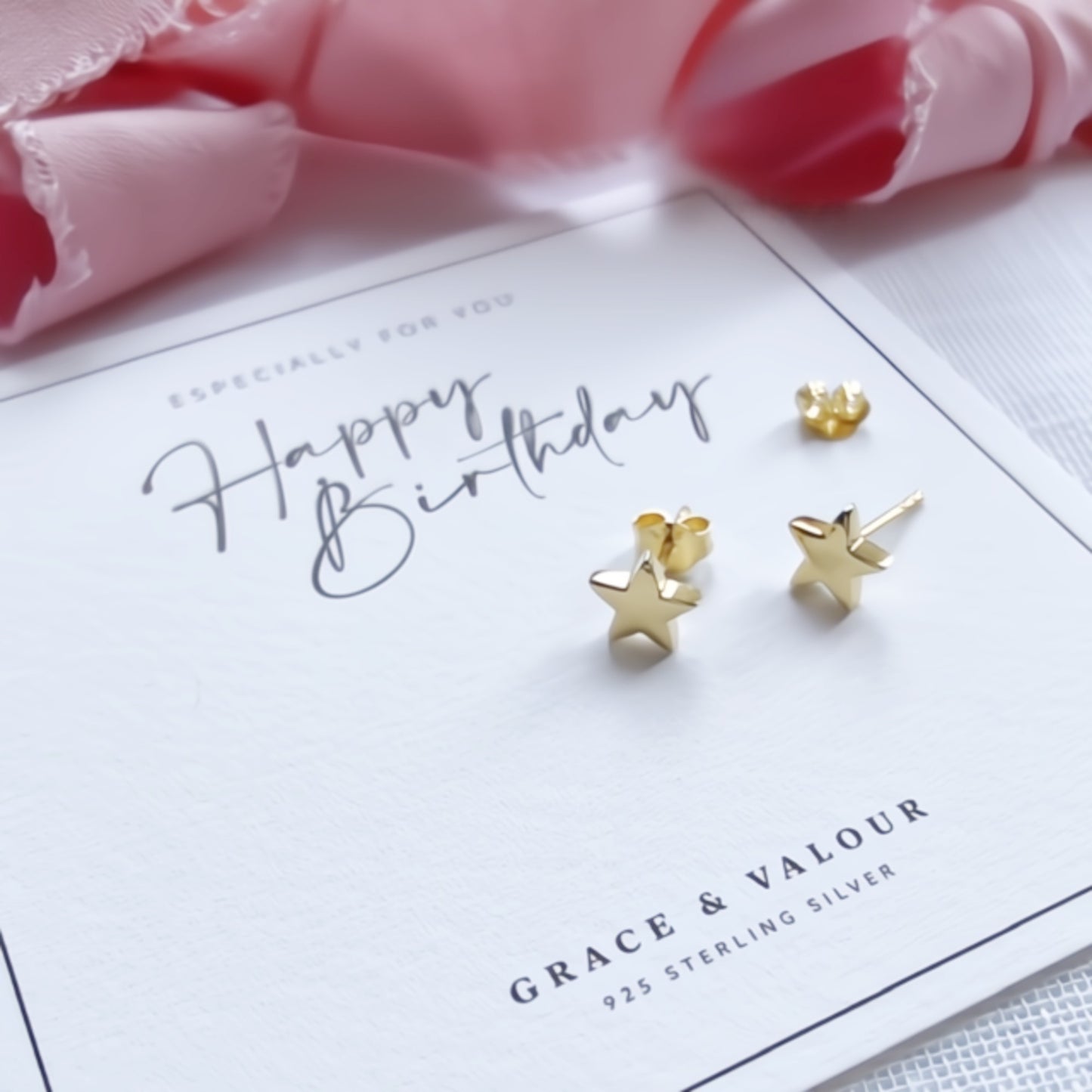 Gold star-shaped stud earrings displayed on a white 'Happy Birthday' greeting card, with pink ribbon in the background. The card shows the brand name Grace & Valour.
