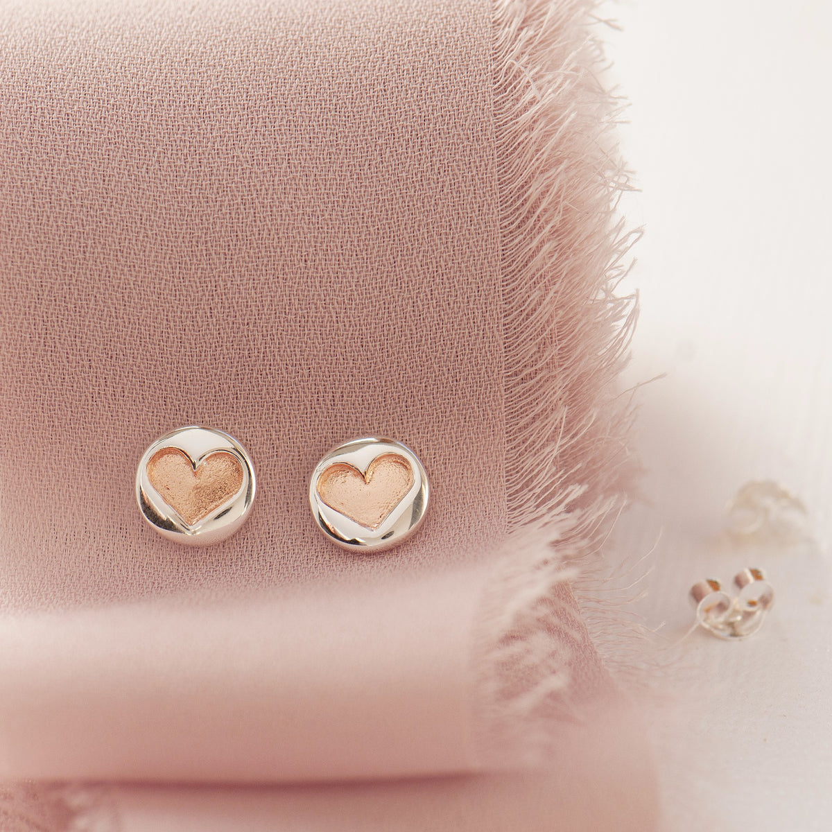 A pair of small heart-shaped stud earrings with polished silver outer halves and rose-gold inner hearts, displayed on a soft pink textured fabric.