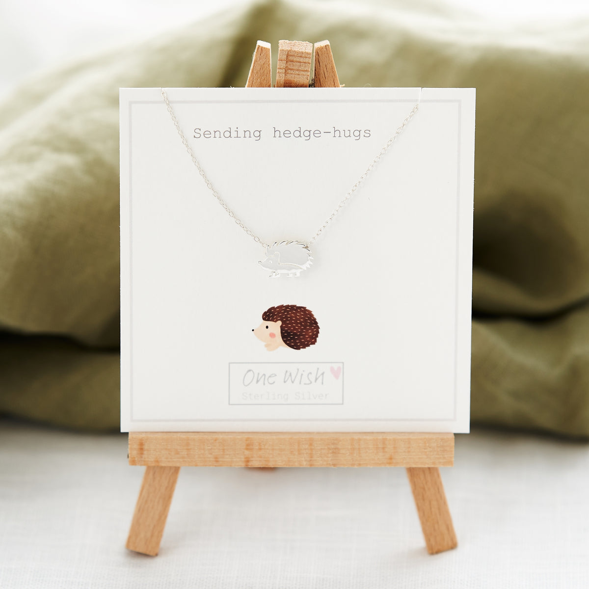 A sterling silver necklace with a hedgehog design on a card that reads 'SENDING YOU HEDGEHUGS', displayed on a wooden easel with a green background.