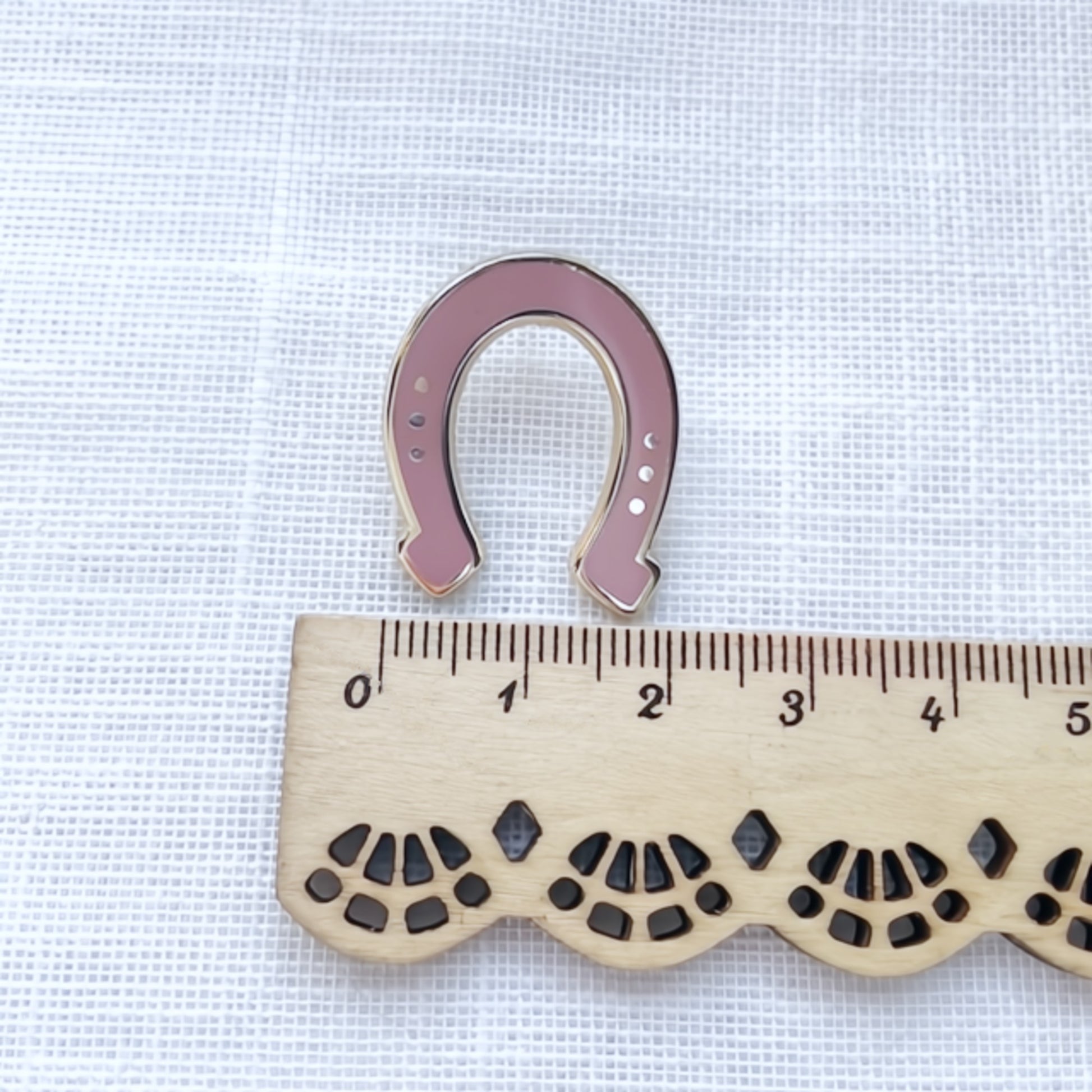 Pink horseshoe enamel pin next to a wooden ruler on a white fabric background