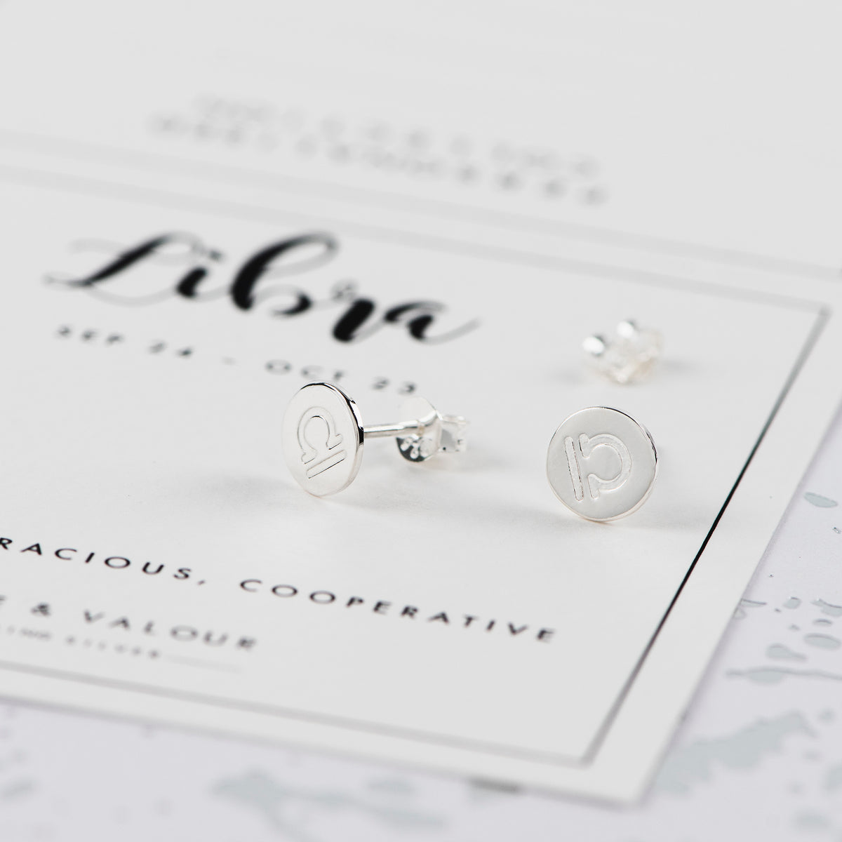 A pair of silver stud earrings with engraved circular symbols, displayed on a white card labeled 'Libra' on a light gray surface.