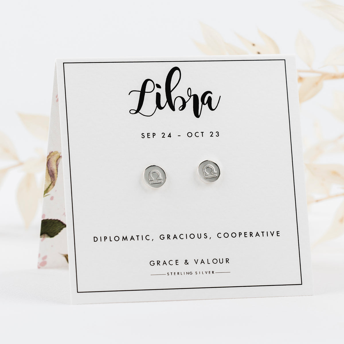 A pair of round sterling silver Libra zodiac stud earrings displayed on a white card backing. The card features black text with the word 'Libra', dates, a personality trait description, the brand 'Grace & Valour', and 'Sterling Silver'.
