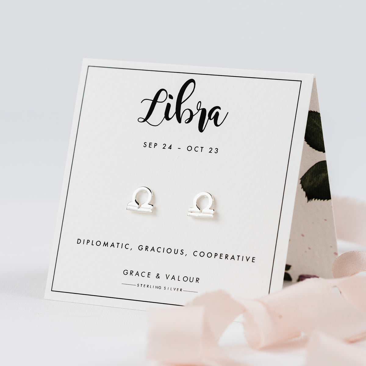 Sterling silver Libra zodiac symbol stud earrings displayed on a white card labeled 'Libra' with decorative pink ribbon in the foreground.