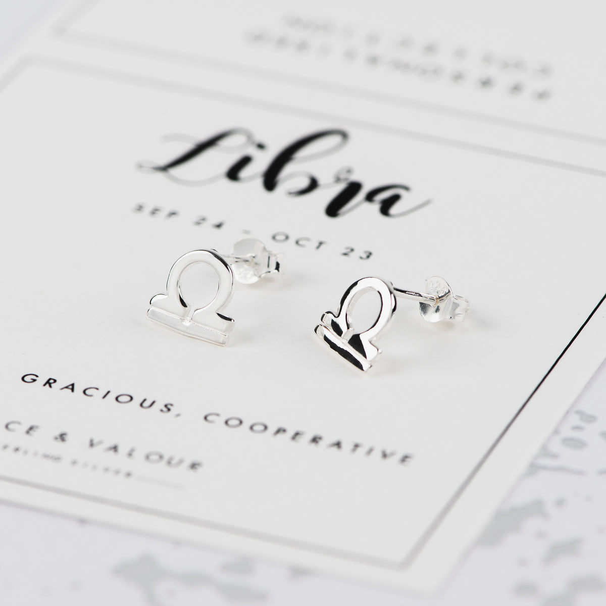 A pair of silver Libra zodiac sign stud earrings displayed on a white card with astrological details, set against a light gray background.