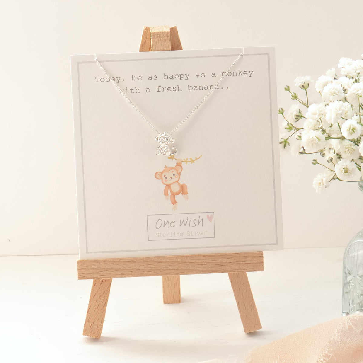 A silver monkey pendant necklace displayed on a white gift card, which is propped up on a small wooden easel. The card features a monkey illustration and the text 'One Wish'.