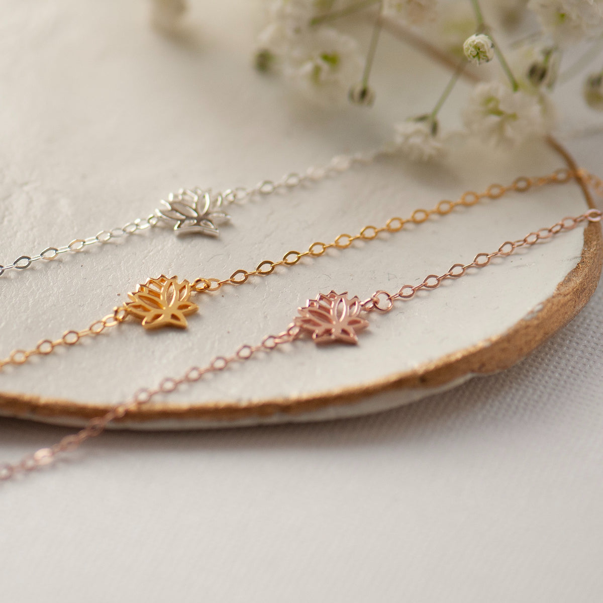 Three delicate chain bracelets with small lotus flower charms in silver, gold, and rose gold, arranged on a round wooden slice with white baby's breath flowers in the background.