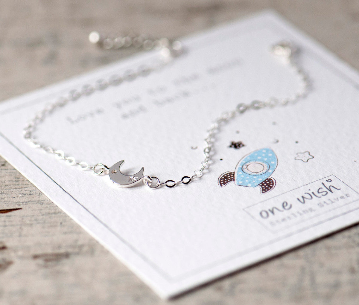 A silver moon-shaped bracelet with a small star charm, displayed on a white card with a personal message that reads 'Love you to the moon and back.