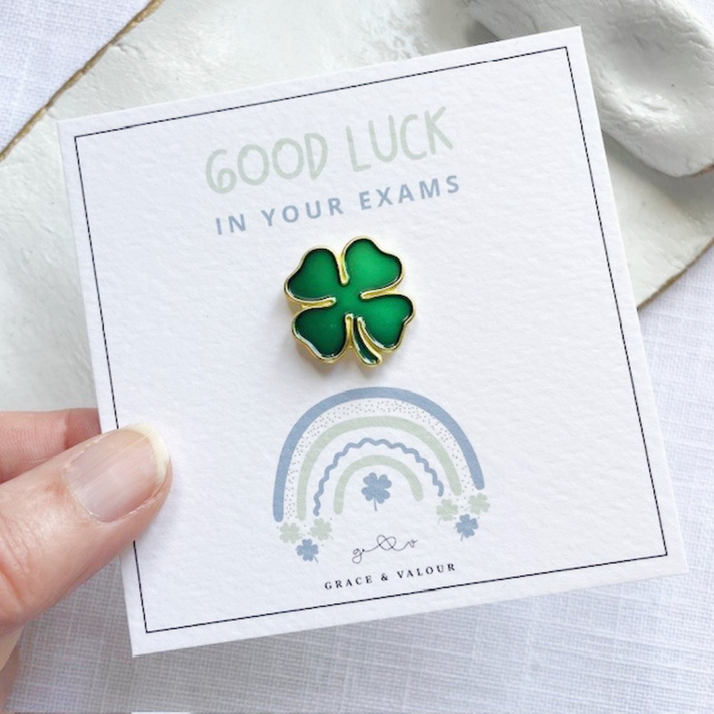 Green four-leaf clover pin on a 'Good Luck in Your Exams' card with a rainbow design.