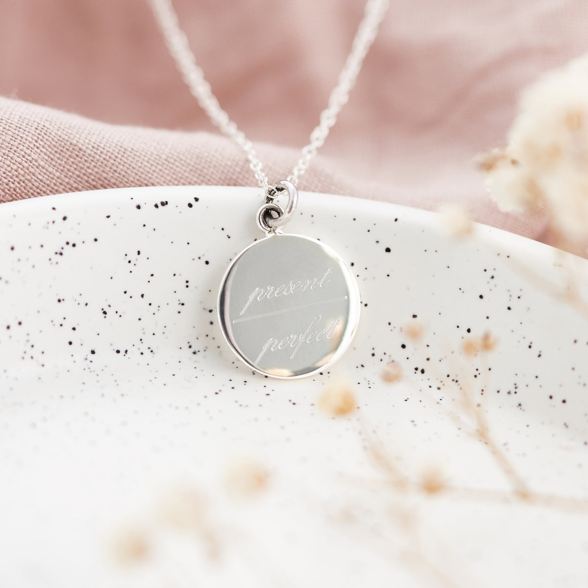 A silver round pendant necklace engraved with 'present future' in cursive, resting on a speckled white ceramic dish with soft pink fabric and dried flowers in the background.