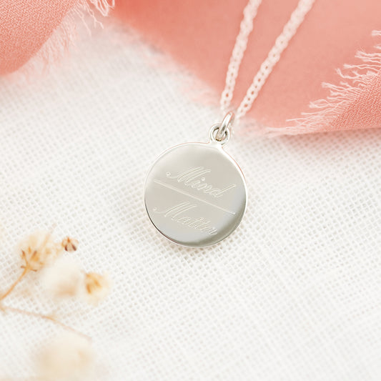 A silver round pendant necklace engraved with the words 'Mind Matter' on a delicate chain, styled on a textured white fabric with soft pink frayed ribbon and dried flowers.