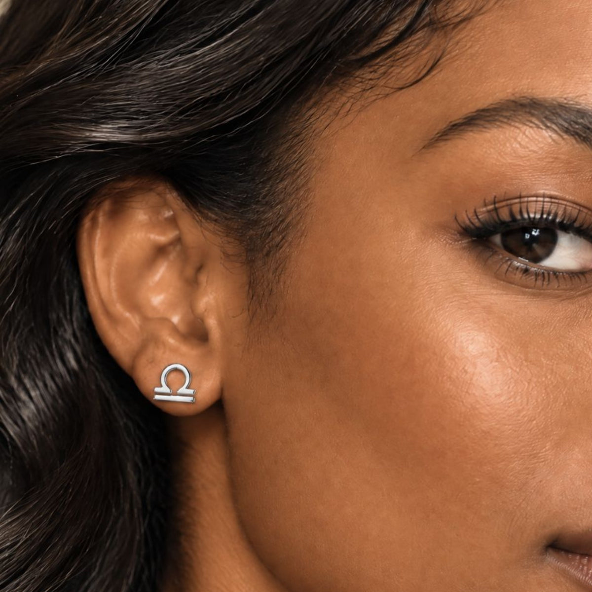 A close-up of a woman's ear wearing a small silver earring with a horoscope symbol, against warm skin tones and dark hair.