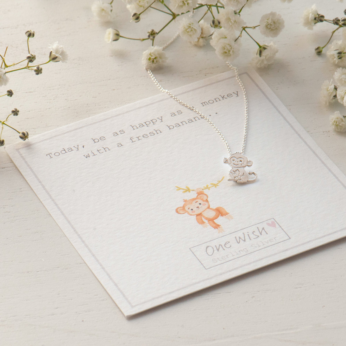 A silver necklace with a monkey pendant is displayed on a white card that reads 'One Wish' with a small orange monkey illustration. The card is styled on a white wooden surface with white baby's breath flowers.